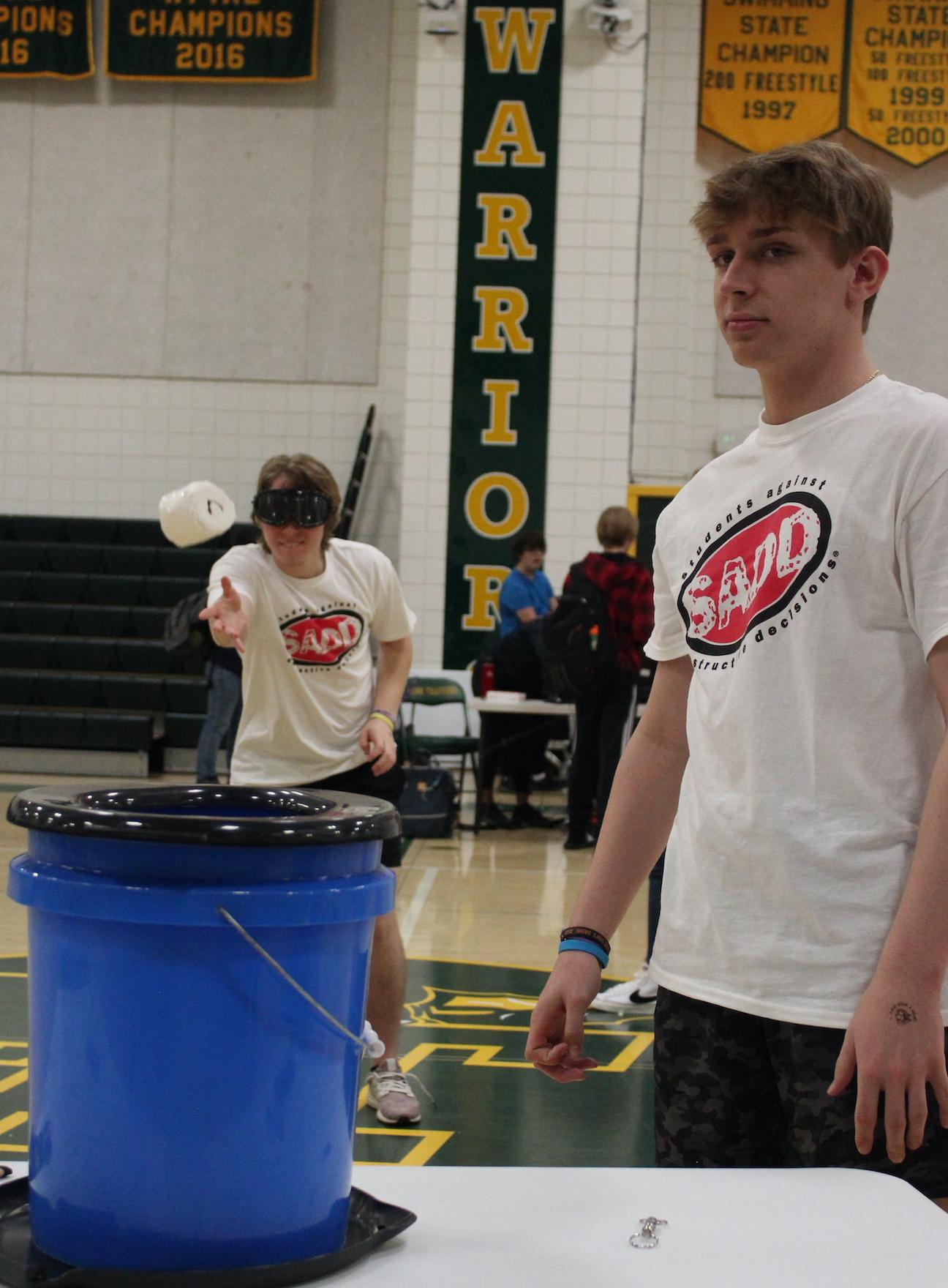 Wearing the simulation goggles, Xavier Solomon tries to toss a roll of toilet paper into a bucket while Nate Loughner observes